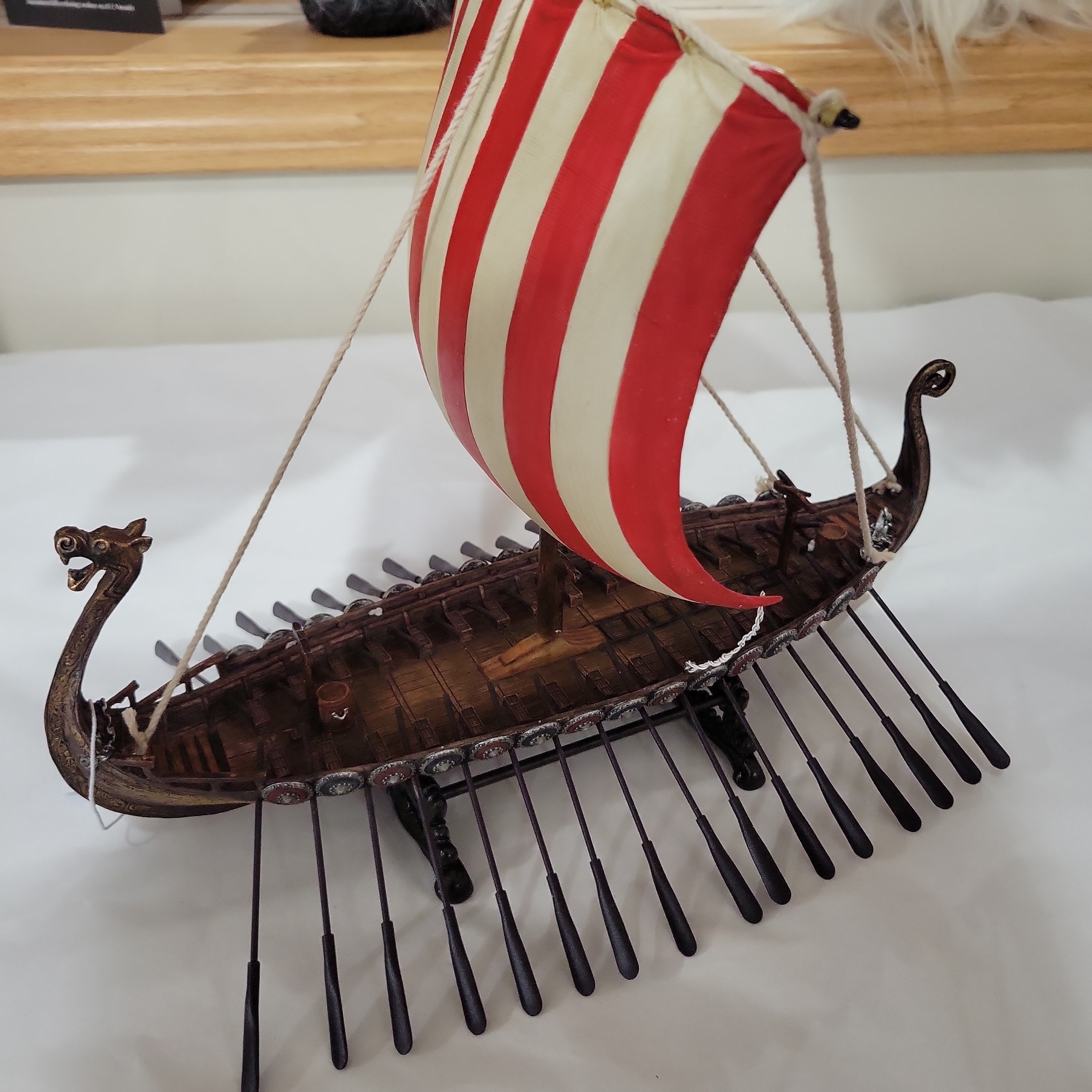 Introducing the Decor: 10" Viking Ship, a meticulously handcrafted model featuring a striped red and white sail, adorned with a dragon head prow and equipped with multiple oars on each side—an ideal piece for Viking enthusiasts.