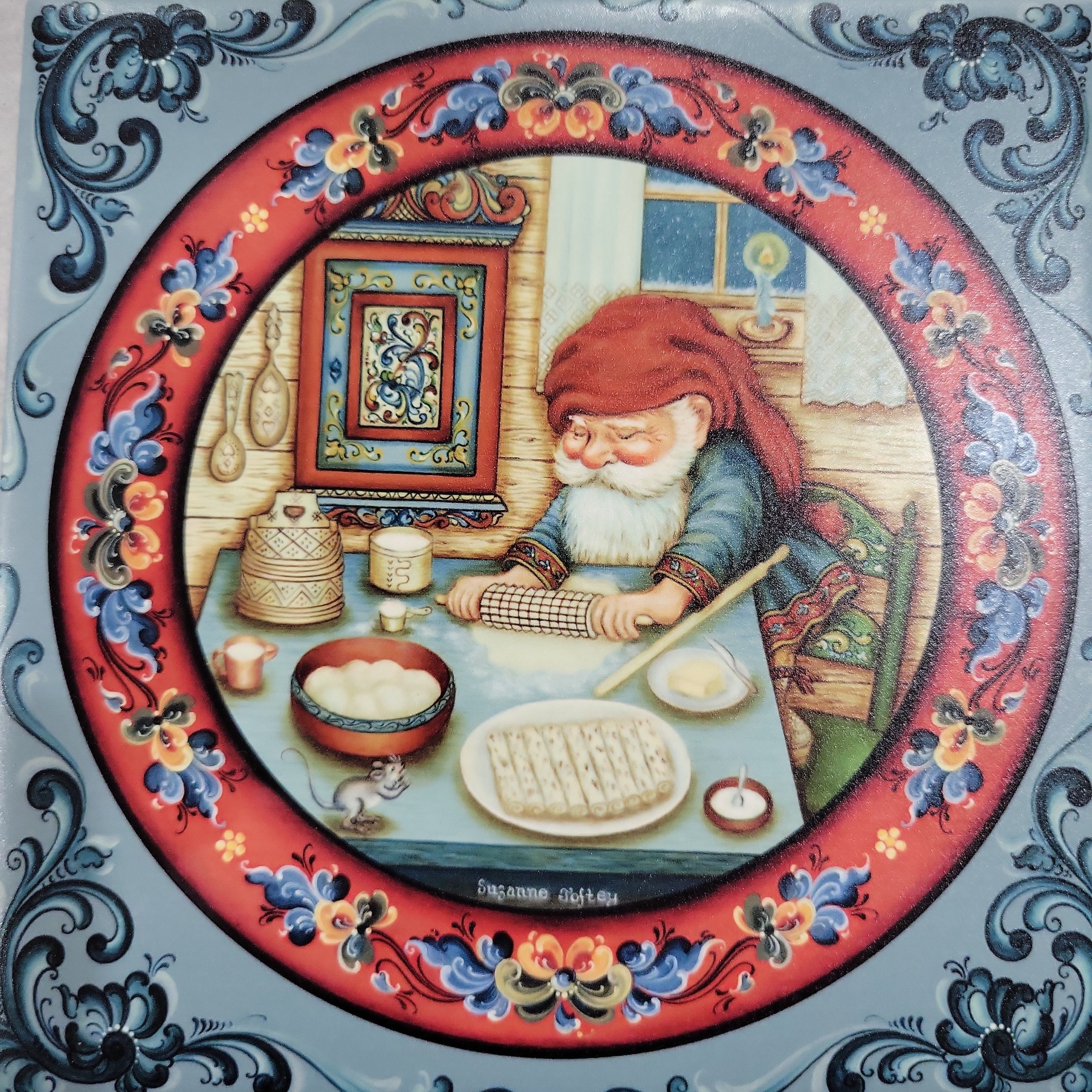 The design portrays a trivet titled "Santa Makes Lefsa 6"," featuring a bearded gnome in a red hat reminiscent of Santa, rolling dough at a table surrounded by baking items. The scene is set within an ornate circular border, making it an ideal addition to your kitchen decor.