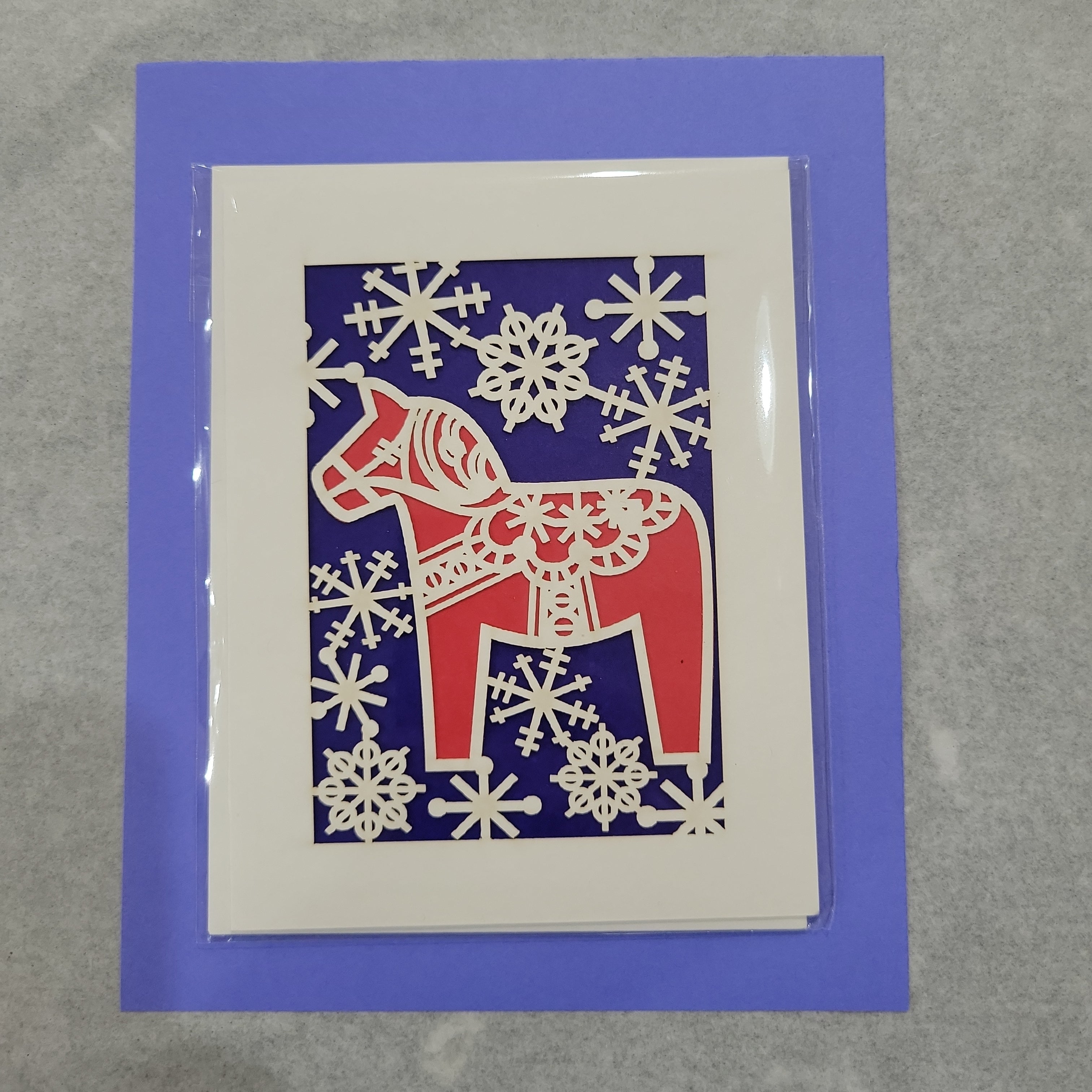 Cards ($3.99): A festive Christmas card with a red horse and white snowflake designs on dark blue, bordered in white, ideal for fans of Danish Christmas carols.