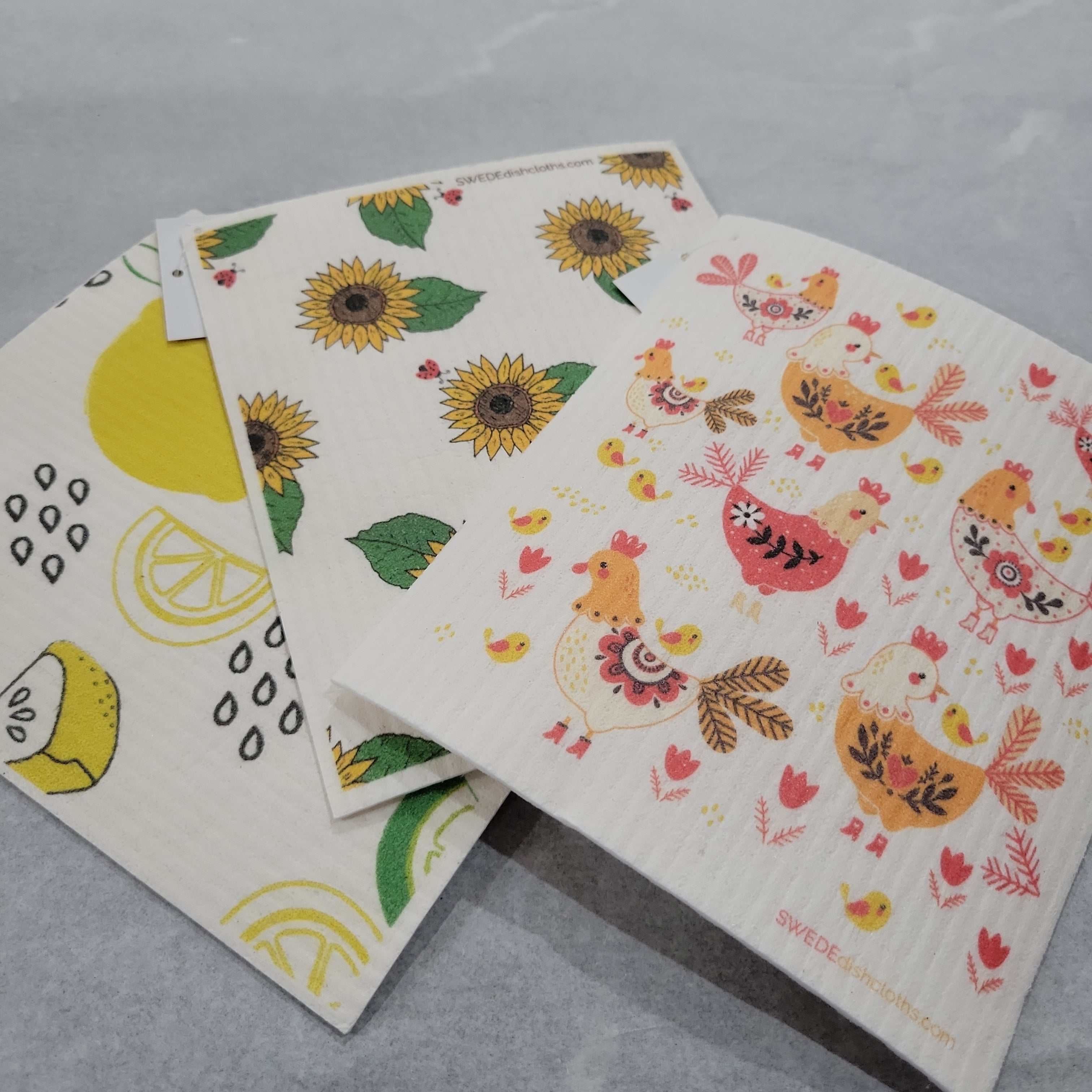 Three eco-friendly Dishcloths are shown on a gray surface, featuring sunflower, lemon, and bird with flower patterns. These super absorbent towels are perfect for your kitchen cleaning needs.
