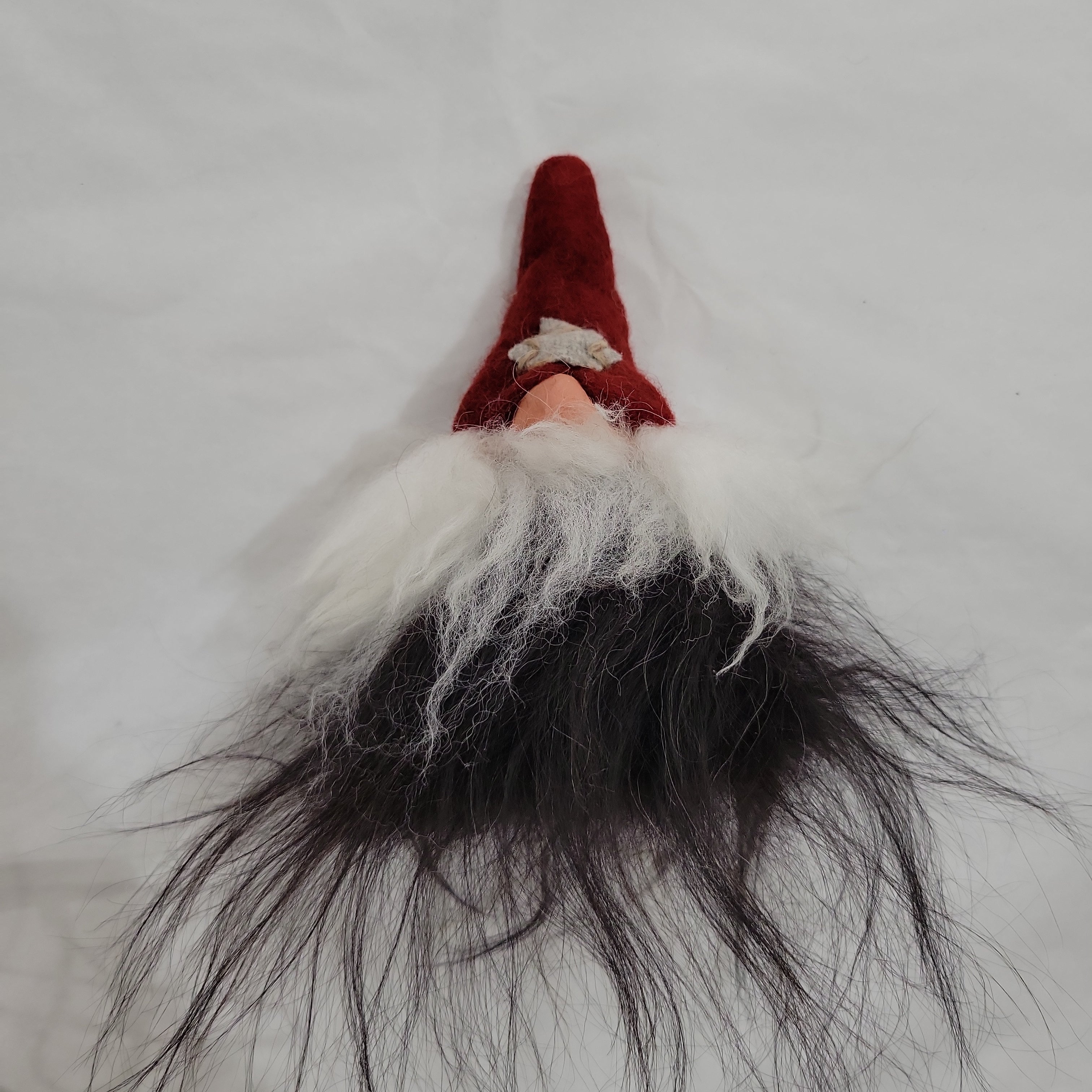 The Gnome: Gumselids Tomte - Gnome 7 is a charming figurine with a red hat and white beard, featuring black sheep wool hair against a white backdrop.