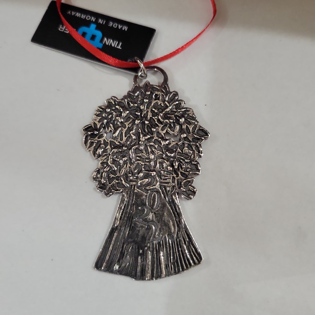 The Ornament: 2022 Pewter Tree Tinn Per is a silver-colored tree-shaped ornament with detailed leaves and trunk, featuring a red ribbon and black tag—ideal for holiday décor.