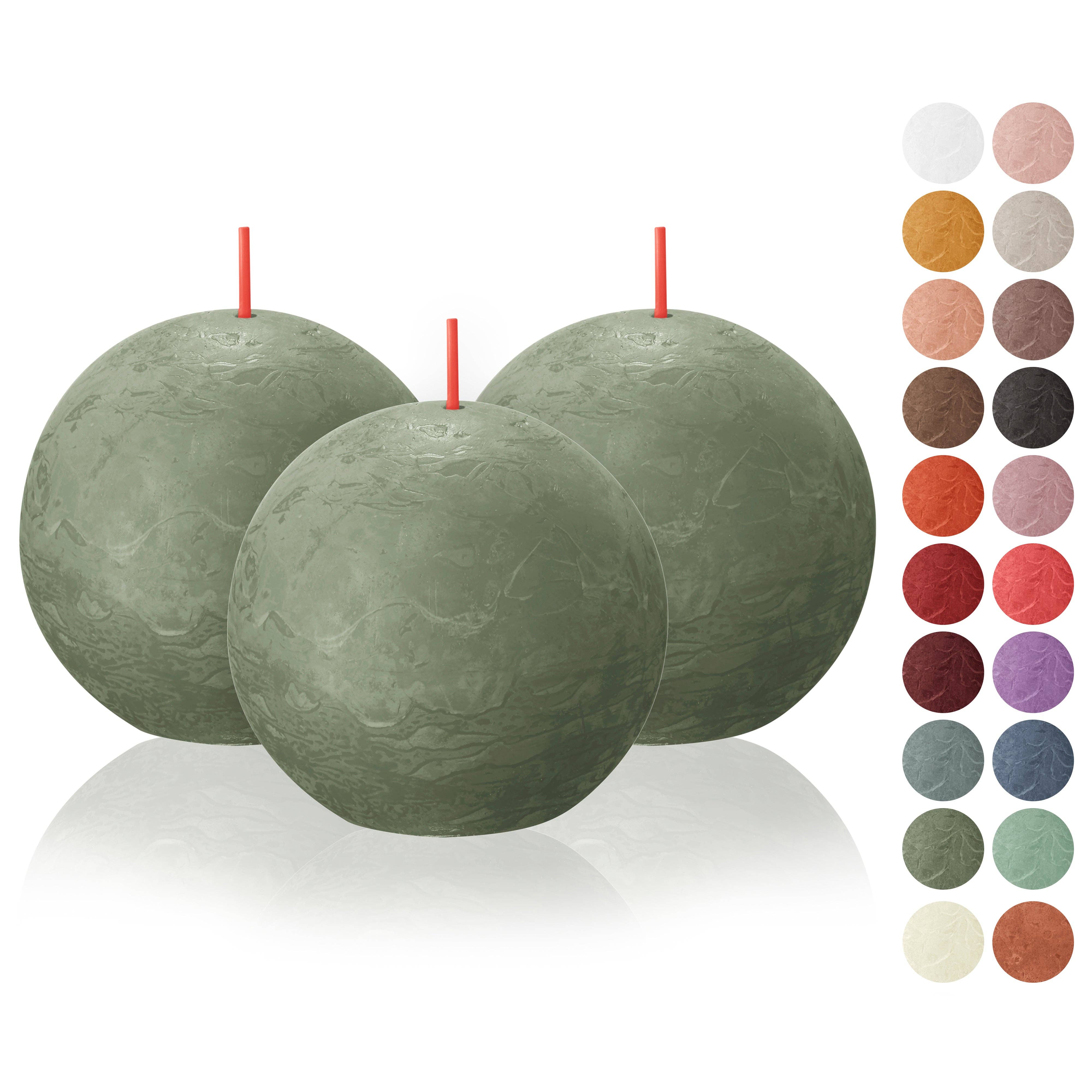 Displayed in front of a color palette with various shades are three Fresh Olive Rustic 3" Ball Candles, distinguished by their large size, green hue, and red wicks. Crafted from plant-based wax, these unscented gems infuse any setting with elegance and eco-conscious style.