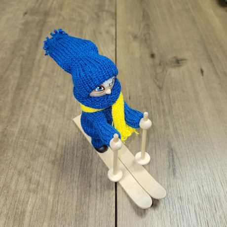 The Swedish Skier Figurine, hand crafted in Sweden, features blue and yellow attire, stands 4.25" tall on 5" long skis, and holds ski poles on a wooden base.