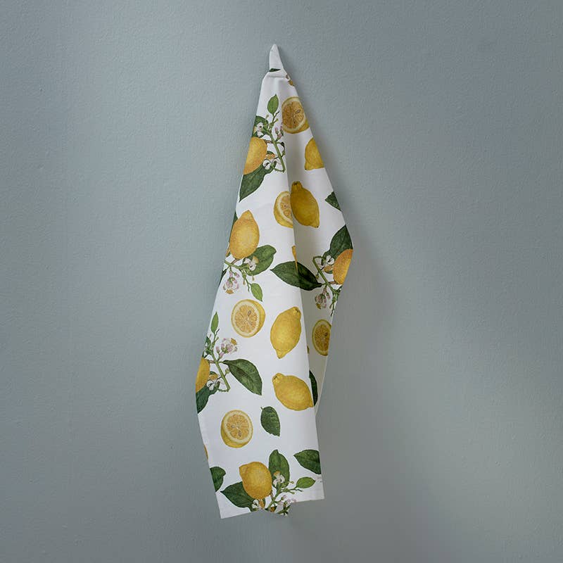 A Tea Towel: Lemons, made from 100% organic cotton and designed by Jim Lyngvild, boasts a delightful lemon and leaf pattern as it hangs gracefully on a pale blue wall.