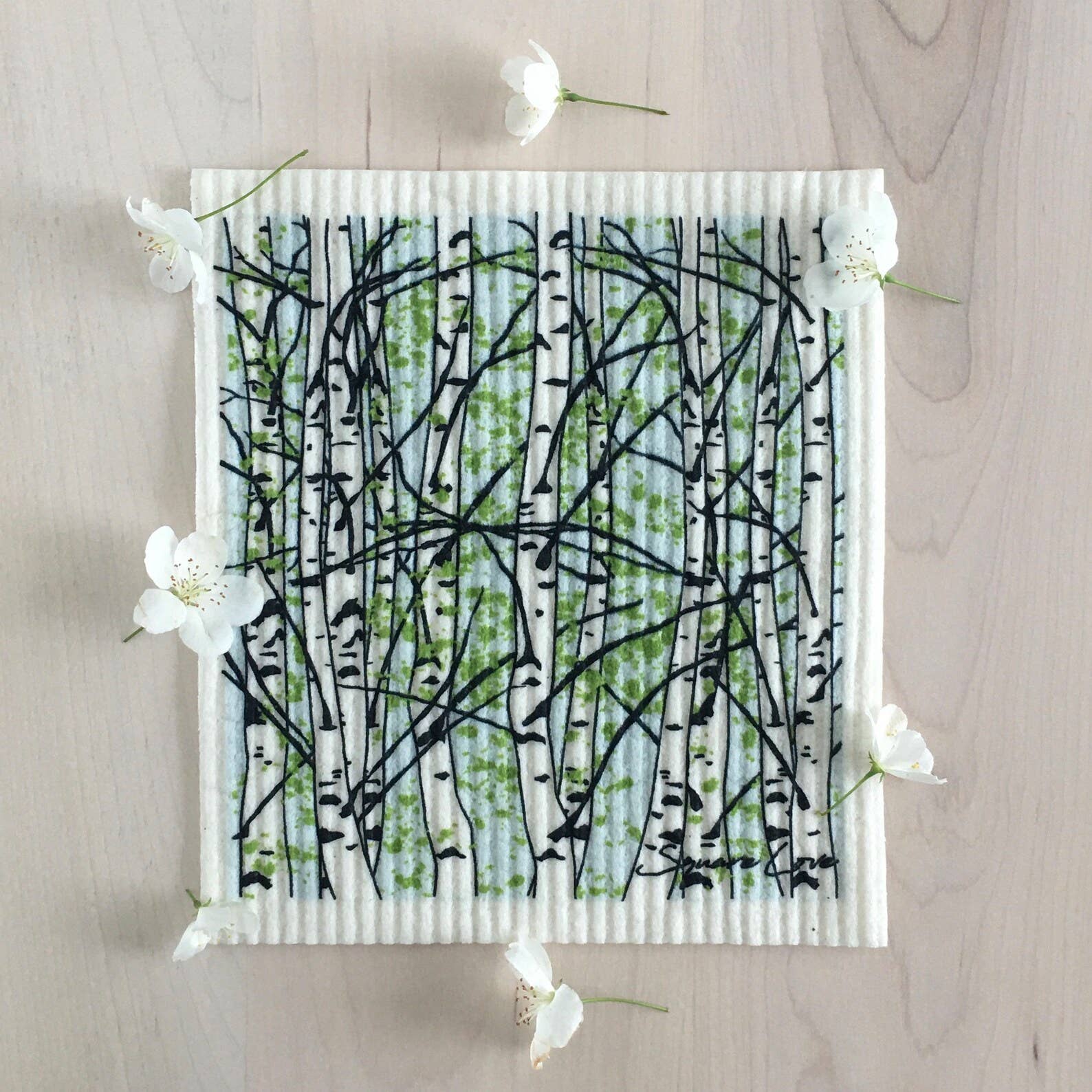 An abstract painting of birch trees with green leaves on a ribbed canvas, accented by small white flowers on a wooden surface, captures the natural charm of the eco-friendly Dish Cloth: Birch Trees Swedish Dishcloth.