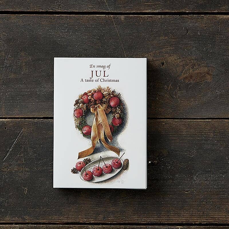 A box of Card Pack: Jul A Taste of Christmas 8 cards sits on a wooden surface, adorned with festive wreaths and cherries. Showcasing delightful Christmas motifs, the packaging is environmentally certified, adding a touch of green to your holiday season.