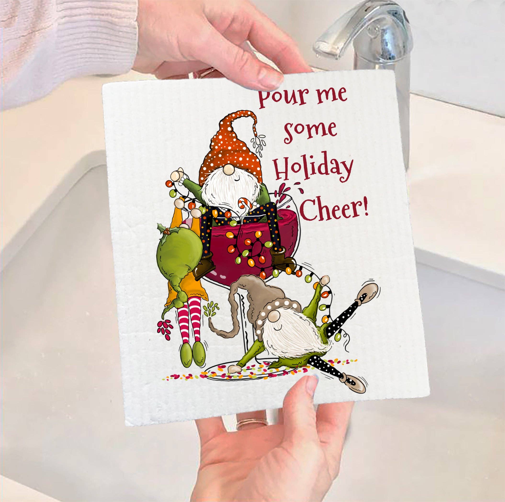 Hands hold a Swedish dishcloth, featuring an eco-friendly design with cartoon elves and the text "Pour me some Holiday Cheer!", near a kitchen sink.