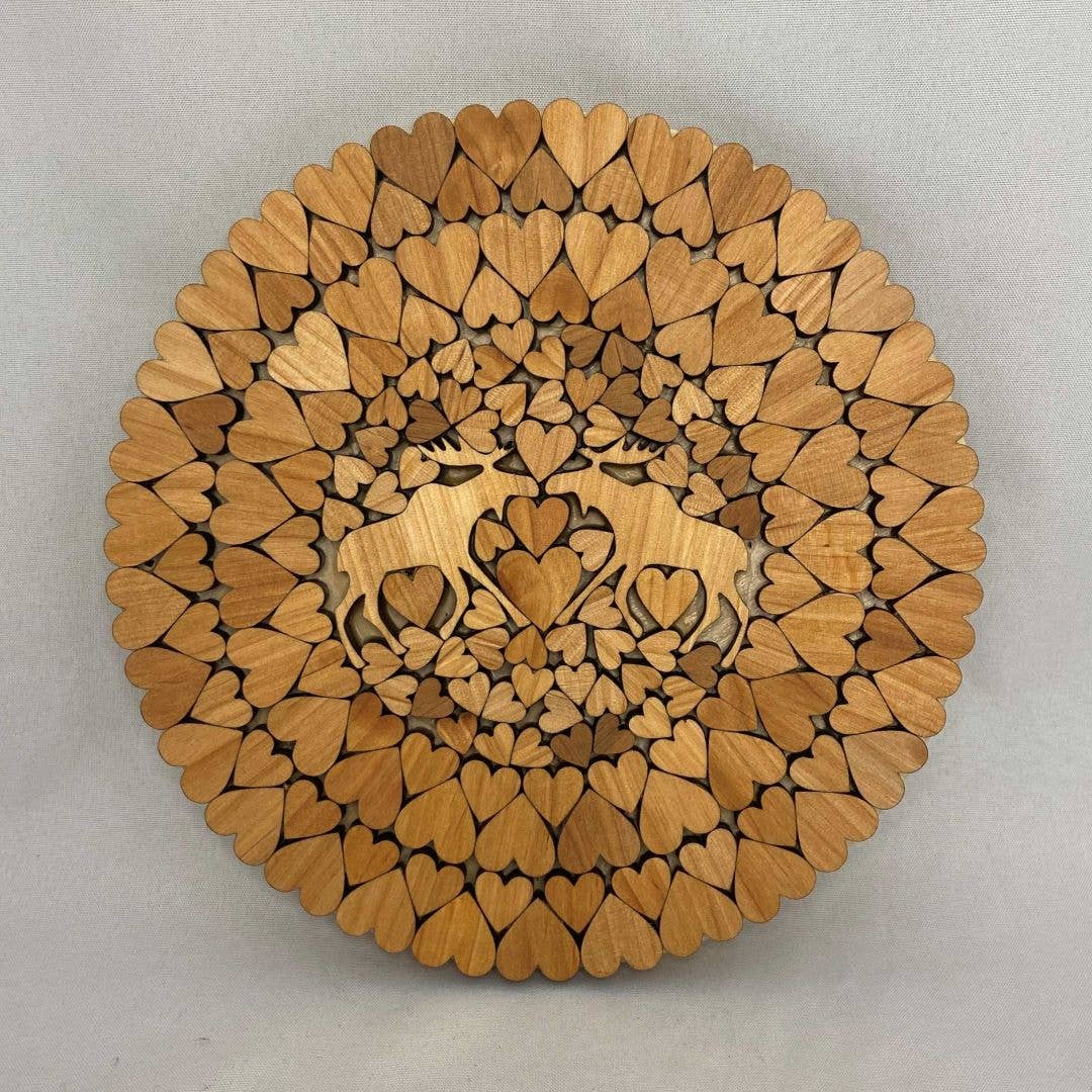 The Two Moose Alder Round Trivet (6" diameter) is a handcrafted wooden trivet with laser-cut hearts in a mandala pattern and two moose facing each other in the center.