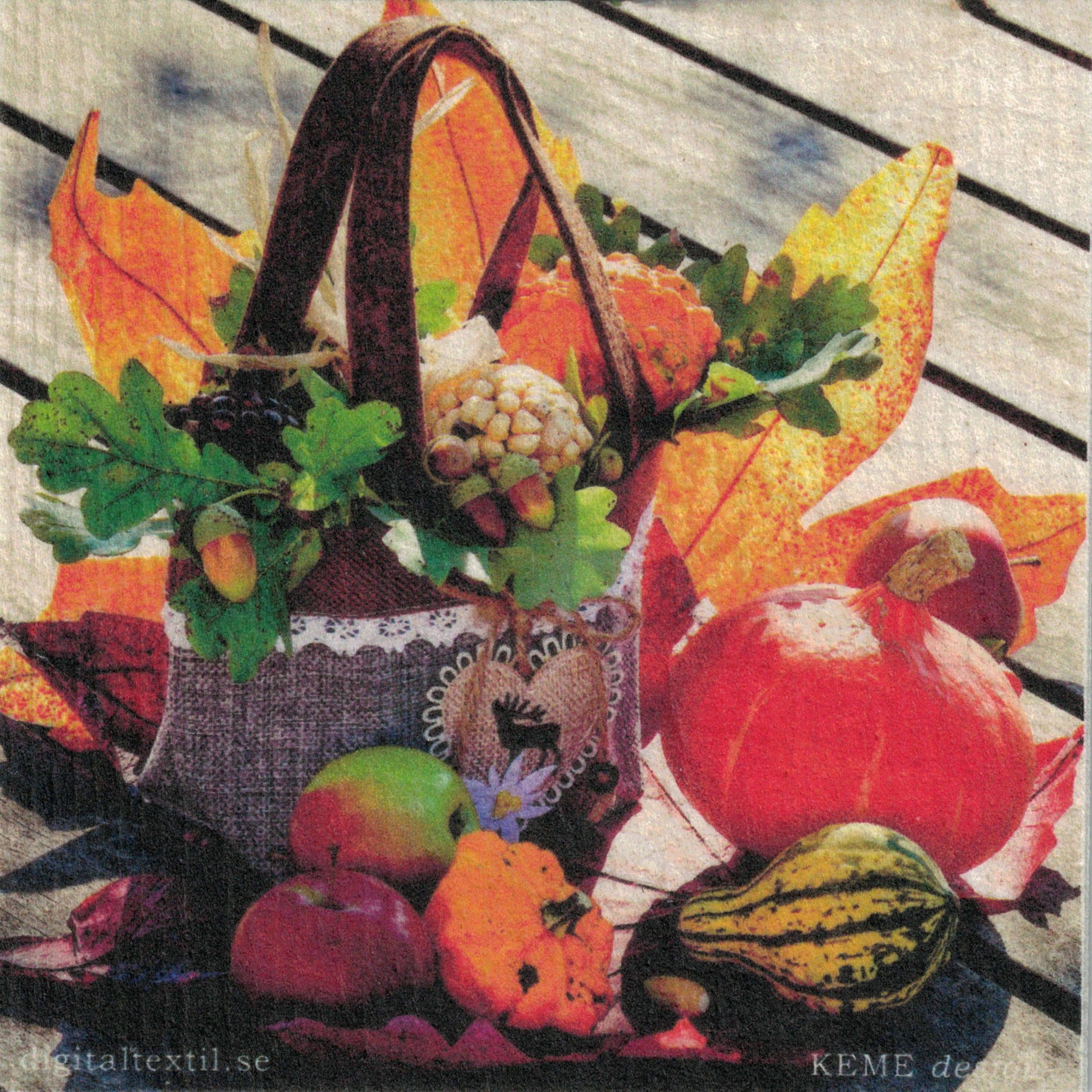 A woven basket overflowing with autumn leaves, corn, and decorative items is surrounded by apples, pumpkins, and gourds on a wooden surface. The Dish Cloth: Fall Harvest adds an eco-friendly touch to the rustic display, perfectly capturing the season's essence.