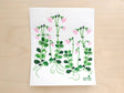 The Linnea Flowers Swedish Dishcloth features a botanical pattern with green vines and pink flowers on a light wooden surface. This eco-friendly, biodegradable dishcloth is highly absorbent, combining sustainability with style for an eco-conscious kitchen choice.