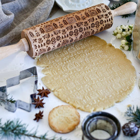 The "Winter Cabin" rolling pin brings Scandinavian design to life with its intricate embossed patterns in the dough, surrounded by cookie cutters, spices, and greenery on a white surface—an ideal choice for those looking for unique handmade gifts.