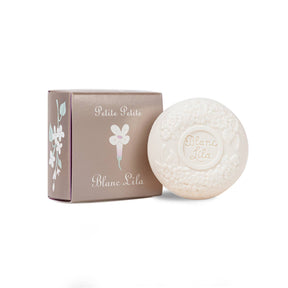 The Petite Petite Blanc Lila Bar Soap is a round embossed lilac soap accompanied by a matching box crafted from recycled packaging, showcasing a floral design and "Petite Petite" text. This cruelty-free soap and its packaging come in soft neutral colors, making it an ideal choice for adding an eco-conscious touch of elegance.
