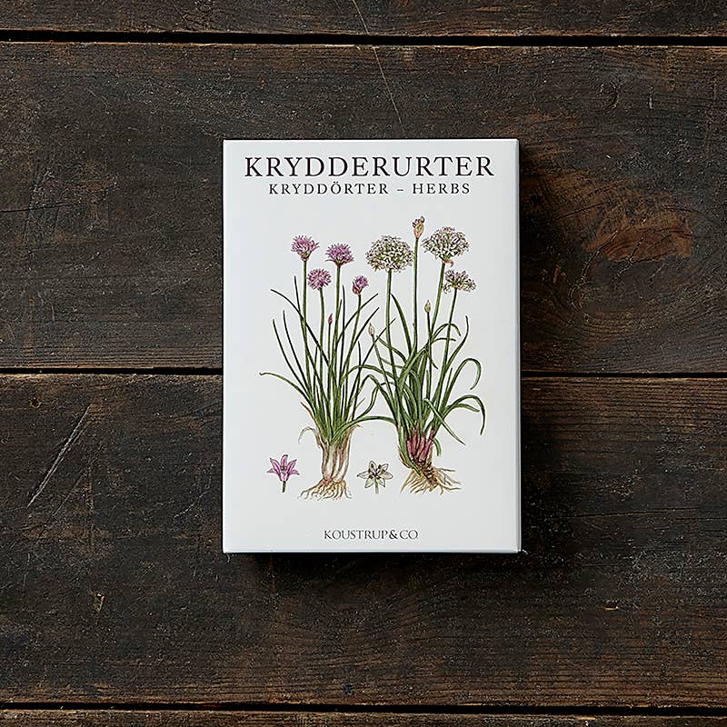 A card pack featuring eight images with envelopes, titled "Herbs," elegantly rests on a wooden surface. Crafted from FSC paper and adorned with vibrant herb motifs along with the text Krydderurter Kryddörter Herbs, it showcases both sustainability and style.