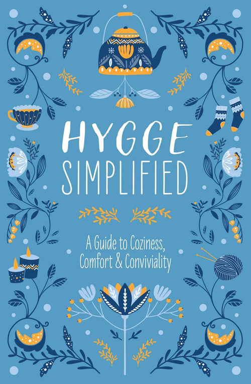 The blue cover of Hygge Simplified: A Guide to Scandinavian Coziness, Comfort, & Conviviality, adorned with a teapot, cup, candles, and floral designs, makes it an ideal housewarming gift.