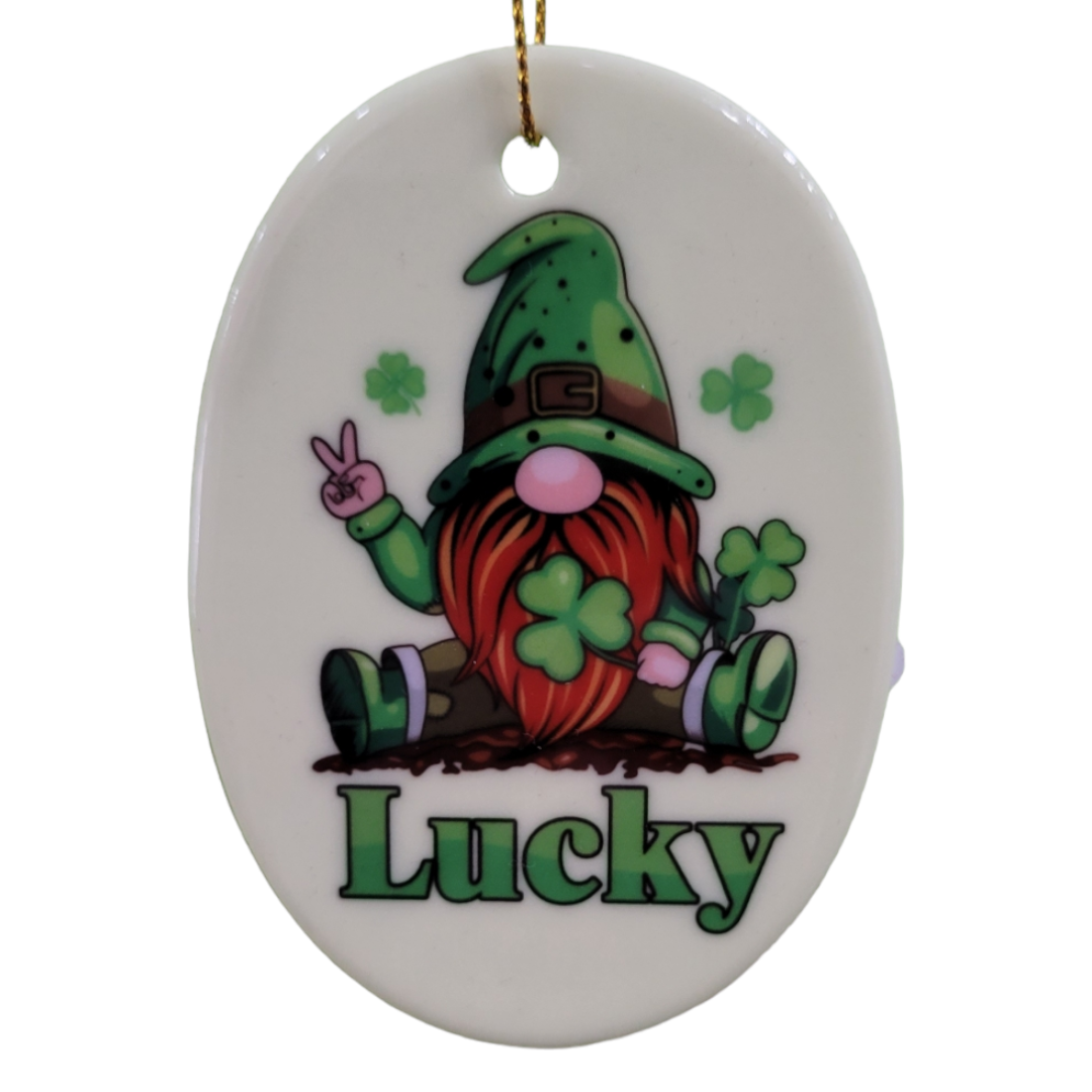 Product Description: The "Lucky" ornament features a gnome with a red beard dressed in green attire, sitting with a peace sign. Shamrocks surround the gnome, and the word "Lucky" is inscribed at the bottom.