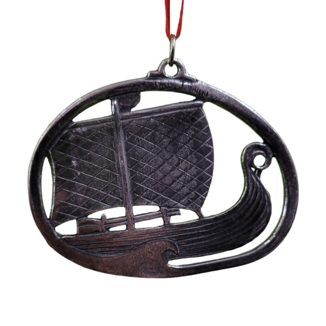 The Ornament: 2" Pewter Viking Ship elegantly hangs from a red ribbon, encapsulating the essence of Norse mythology. Encircled within an oval frame, this exquisite piece of holiday décor infuses your space with historical charm.