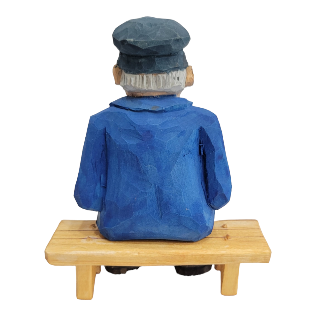 Hand-painted wood carved figurine titled "Elmer Axelson" by Bill Erickson, depicting a man with a cap and blue coat sitting on a bench, viewed from the back.