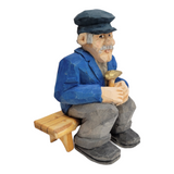 The "Elmer Axelson" figurine by Bill Erickson features an elderly man sitting on a bench, dressed in a blue jacket and cap, holding a small brown pipe. Hand-painted and intricately carved from wood, this piece beautifully captures timeless charm and detail.