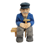 The "Elmer Axelson" figurine by Bill Erickson is a hand-painted, wood-carved representation of an older man. He is depicted in a blue jacket and cap, with a gentle smile, holding a cane while sitting comfortably on a wooden bench.