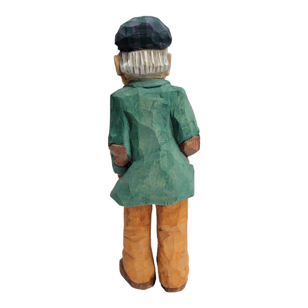 This 8 1/2 inch wood carved figure, titled "Man w/Pipe" by Bill Erickson, features a person with a green shirt, orange pants, black cap, and long white hair, viewed from the back.