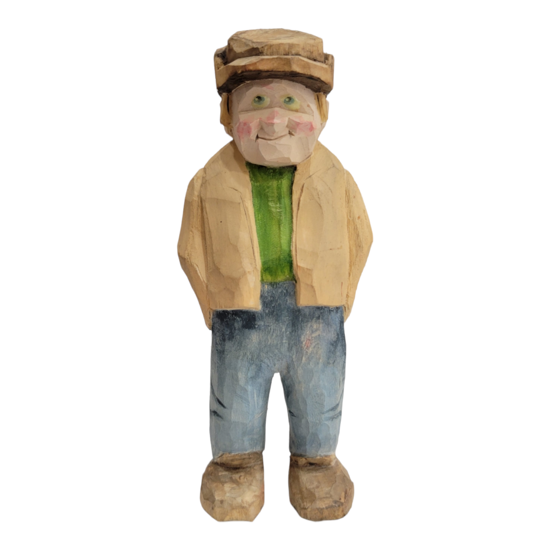 The 6 1/2-inch "Sven" wood carved figurine by AM portrays a person in a hat, green shirt, tan jacket, and blue pants. This delightful sculpture captures a joyful expression with its hands playfully positioned behind its back.