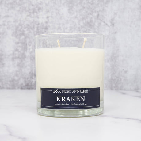 An 8oz Fjord & Fable Kraken candle in a clear glass jar, featuring amber, sea salt, driftwood, and leather notes, sits on a white surface with a gray background.
