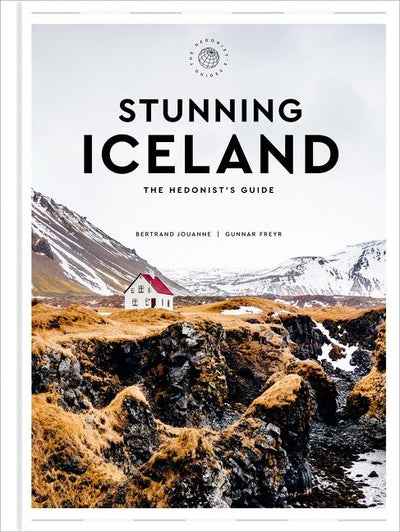 Cover image of the book *Stunning Iceland The Hedonist’s Guide*, featuring breathtaking photography of a house nestled in rocky terrain with snowy mountains, creating a captivating scene for an unforgettable Iceland travel guide.
