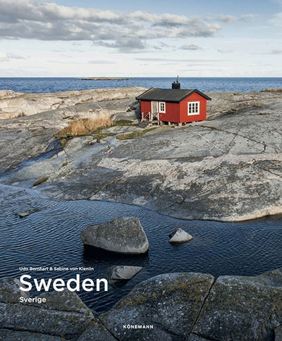 Nestled on Swedens rocky seashore, this small falun-red wooden cabin embodies a Scandinavian idyll beneath a cloudy sky, as depicted in the book Sweden (Spectacular Places).