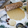 A Birds & Flowers Embossing Rolling Pin elegantly sits on intricately patterned cookie dough. Surrounding it are cookie cutters, spices, and greenery on a flour-dusted surface, capturing the essence of Scandinavian design—a delightful addition for crafting handmade gifts.