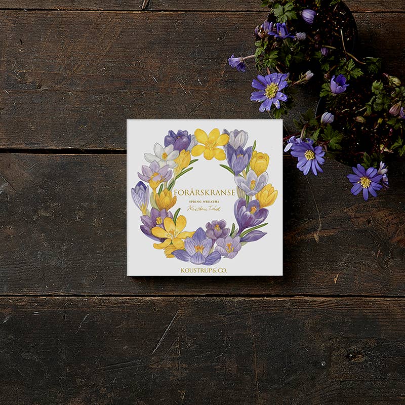 A square card folder labeled "Card Pack: Spring Wreaths - Square Card Folder 8 Cards" sits elegantly on a dark wooden surface, adorned with a touch of gold printing, next to a small bouquet of purple flowers.