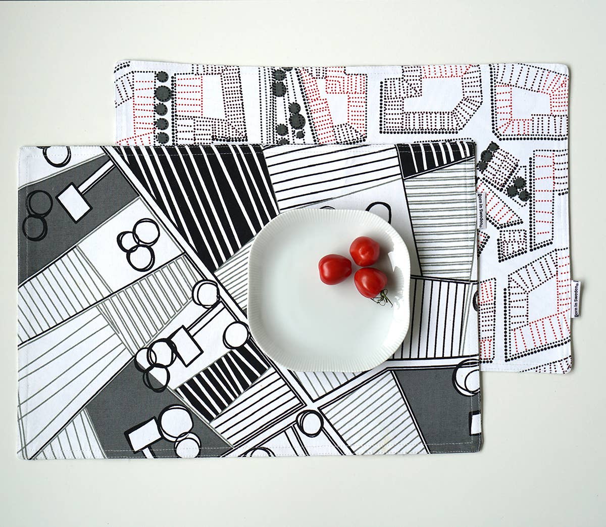 A white plate with three cherry tomatoes is set on a stylish, 47cm x 34cm double-sided cotton placemat from Born in Sweden, featuring black, white, and red patterns.